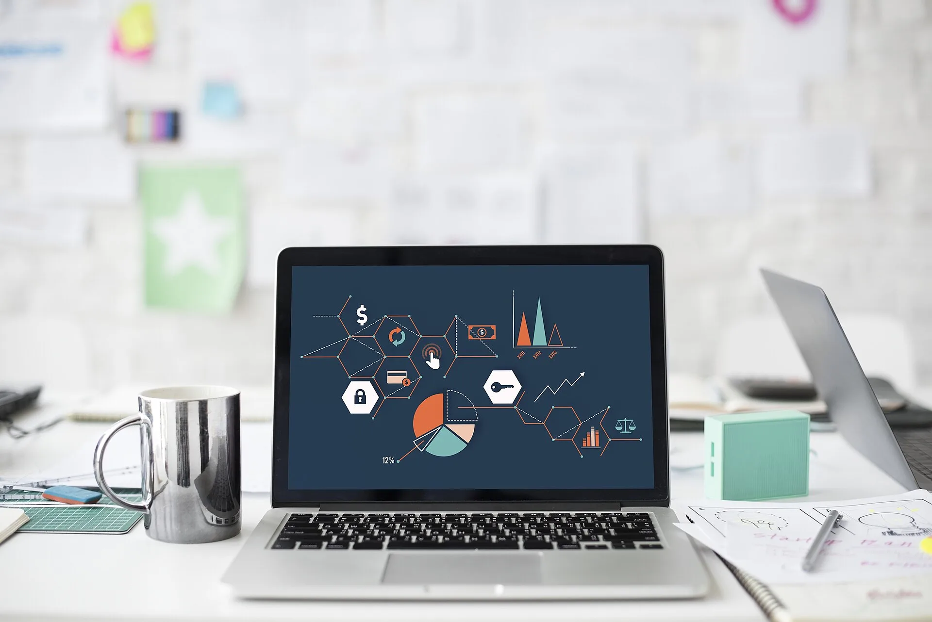 A laptop (MacBook) open on a cluttered desk, displaying a screen with many charts on it. The background has a wall with many papers pinned to it.
