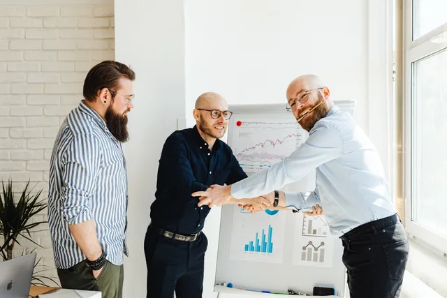 Three businessmen engage in a lively discussion around a whiteboard with charts in a sunlit office. Customer success software launch review explained for UK teams