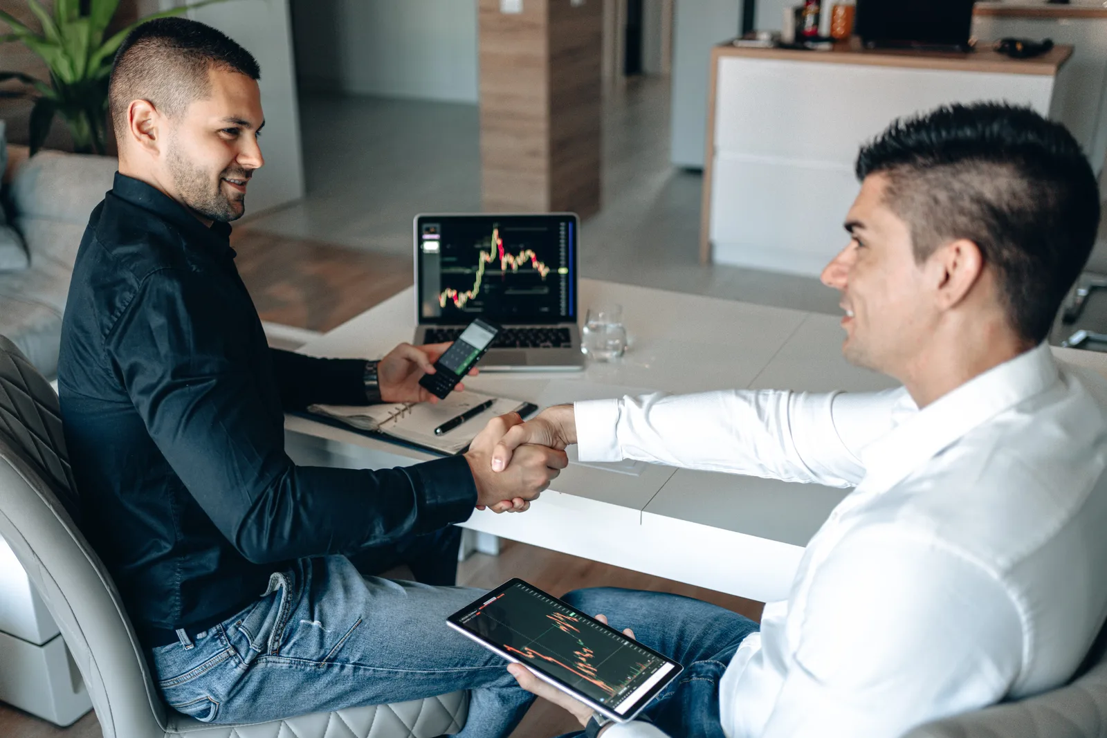 Two businessmen shake hands over a successful stock trading agreement using technology in a modern office.