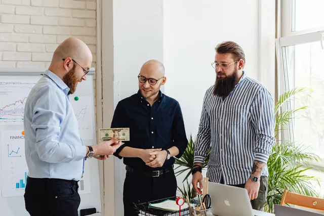 Three businessmen exchanging money in a modern office with financial charts. How customer success software pricing models affect budgets